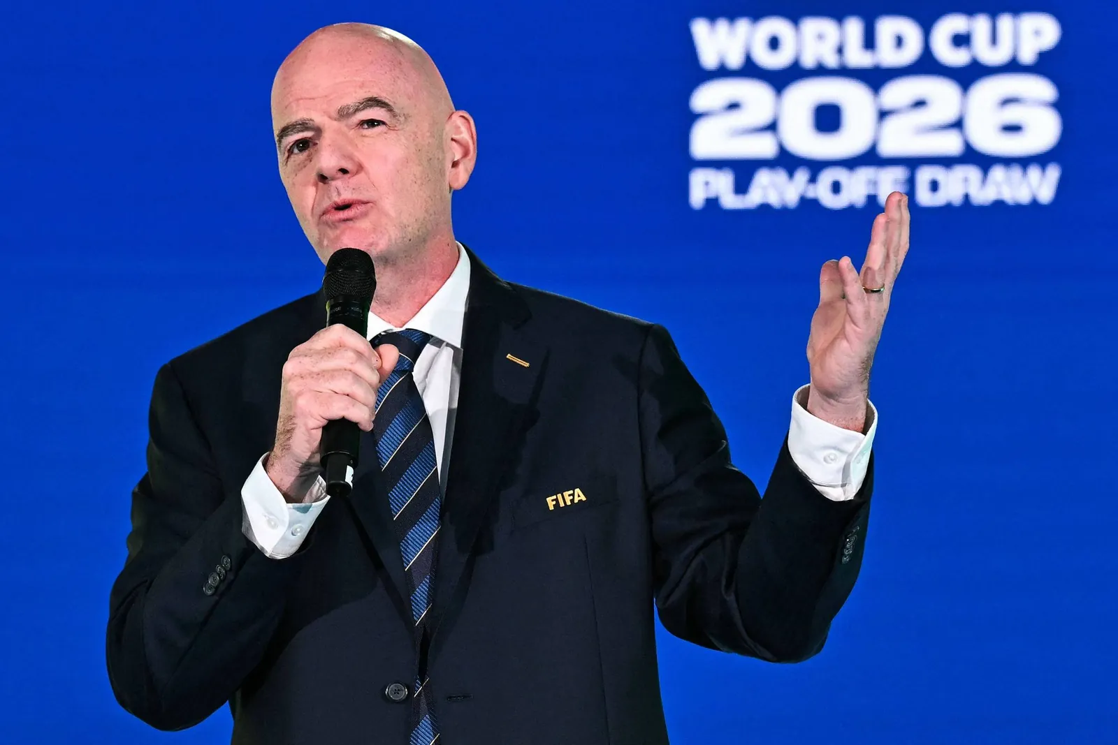 Homem de terno escuro com gravata listrada segura microfone e gesticula com a mÃ£o direita. Fundo azul exibe texto 'WORLD CUP 2026 PLAY-OFF DRAW'.